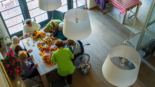 EIne Gruppe älterer Leute mit Betreuern sitzt um einen Tisch herum und bastelt mit Blumen.