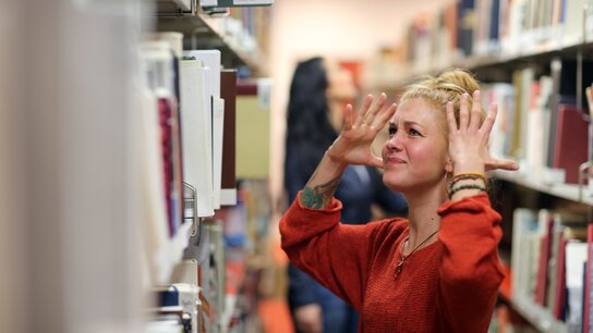 Eine blonde Frau steht zwischen zwei Regalen in einer Bücherei mit einem genervten Gesichtsausdruck und hat die Hände ärgerlich Richtung Kopf gestreckt.