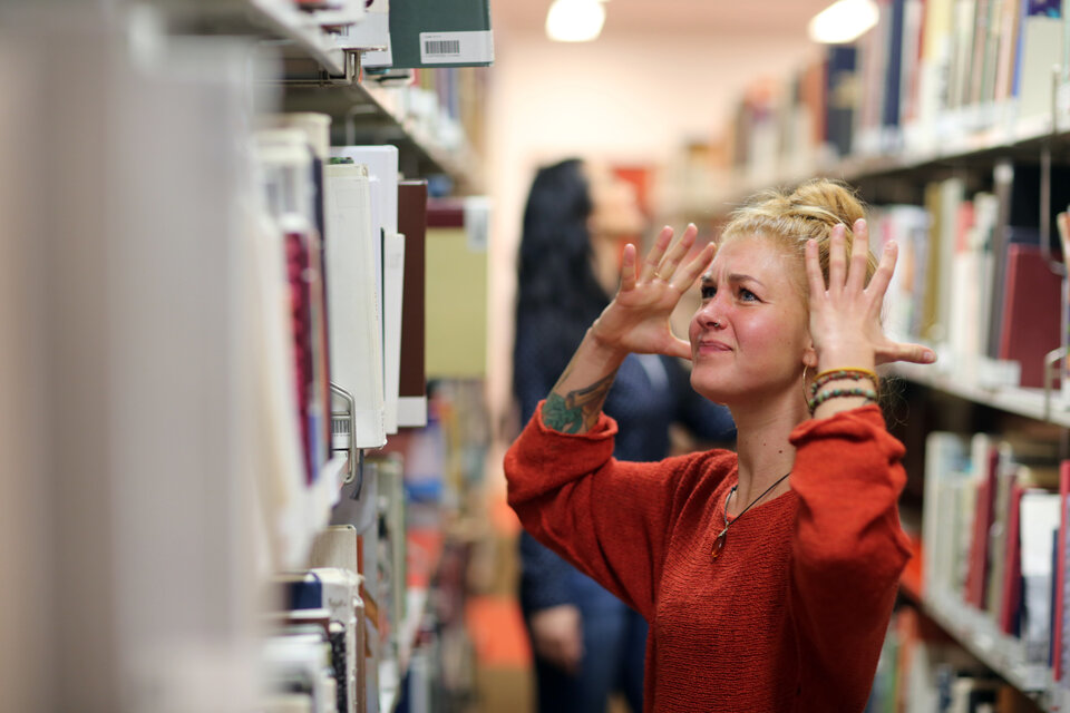 Eine blonde Frau steht zwischen zwei Regalen in einer Bücherei mit einem genervten Gesichtsausdruck und hat die Hände ärgerlich Richtung Kopf gestreckt.