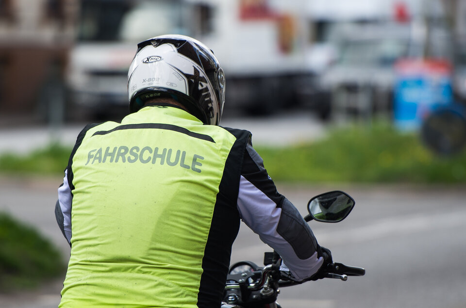 Motorradfahrer mit Sicherheitsweste "FAHRSCHULE" auf einer Straße, im Hintergrund verschwommene Fahrzeuge.