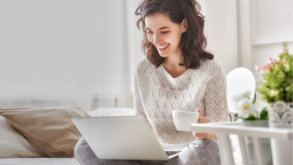 Lächelnde Frau sitzt auf dem Sofa mit Laptop und Tasse in der Hand, umgeben von Pflanzen und hellem Licht.