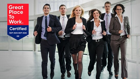 Sechs lächelnde Personen in formeller Kleidung gehen gemeinsam in einem modernen Bürogebäude. Links das Logo "Great Place To Work".