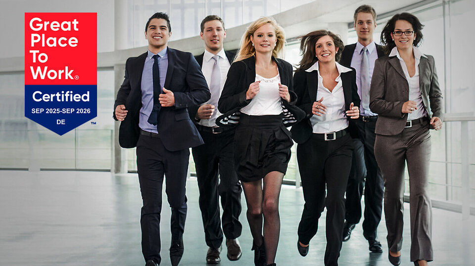 Sechs lächelnde Personen in formeller Kleidung gehen gemeinsam in einem modernen Bürogebäude. Links das Logo "Great Place To Work".