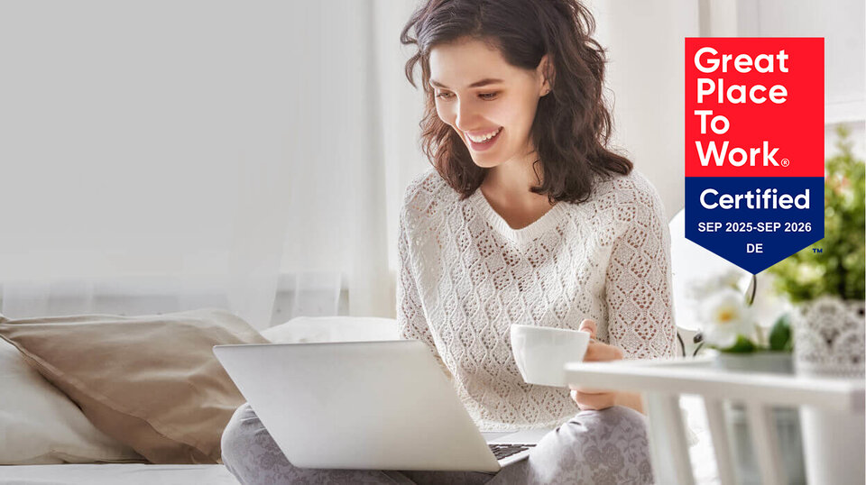 Lächelnde Frau sitzt mit Laptop und Tasse in einem hellen Raum, umgeben von Pflanzen, mit Auszeichnung für Arbeitsplatzqualität.