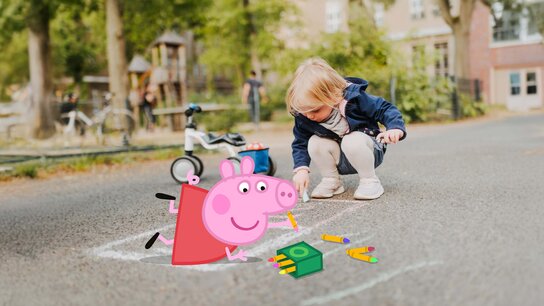 EIn kleines Mädchen und Peppa malen mit Straßenmalkreide. Neben dem Mädchen steht ein Laufrad und im Hintergrund ist ein Spielplazt zu sehen.