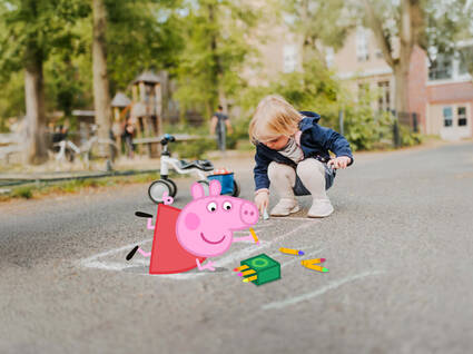 EIn kleines Mädchen und Peppa malen mit Straßenmalkreide. Neben dem Mädchen steht ein Laufrad und im Hintergrund ist ein Spielplazt zu sehen.