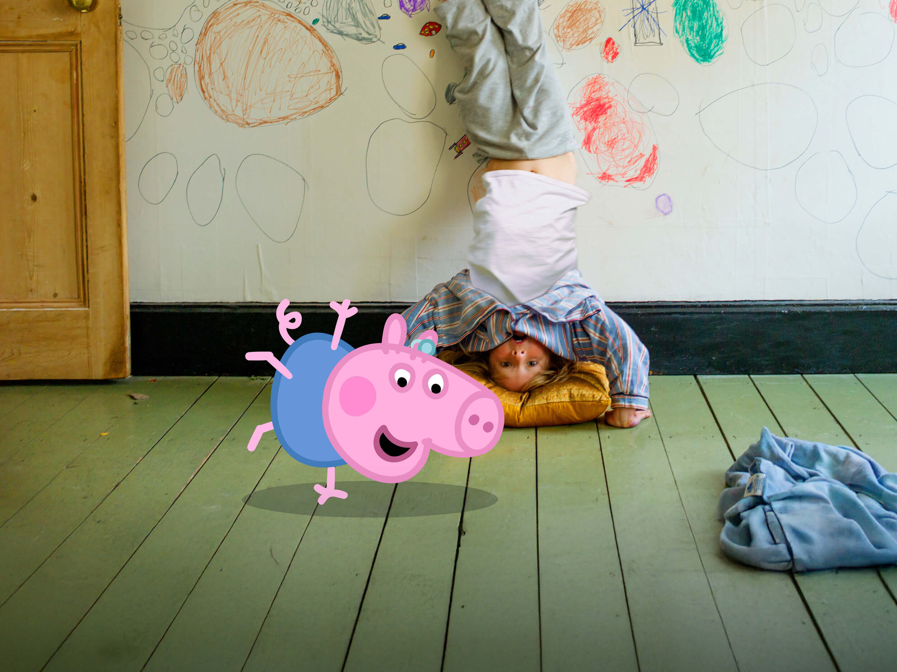 Ein Junge macht an einer bemalten Kinderzimmerwand einen Kopfstand auf einem Kissen. Peppa turnt davor herum.