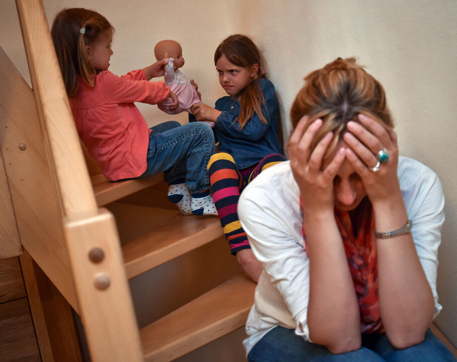 Zwei Kinder spielen auf einer Treppe, während eine Erwachsene frustriert mit den Händen im Gesicht sitzt.