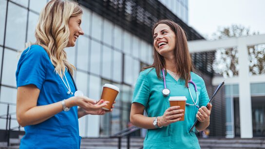 Zwei Ärztinnen stehen mit jeweils einem Kaffee Becher in der Hand vor einem Krankenhaus und unterhalten sich fröhlich.