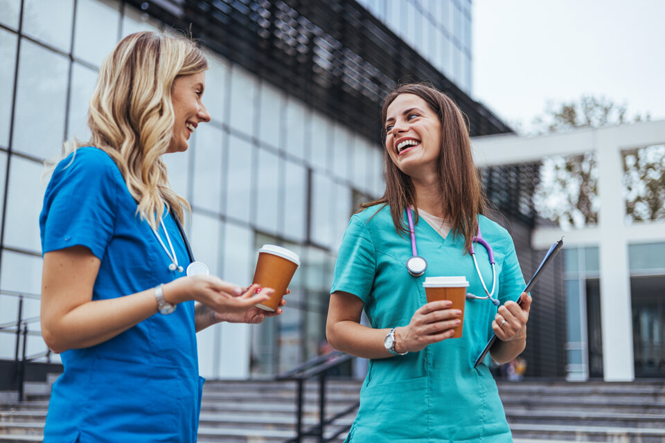 Zwei Ärztinnen stehen mit jeweils einem Kaffee Becher in der Hand vor einem Krankenhaus und unterhalten sich fröhlich.