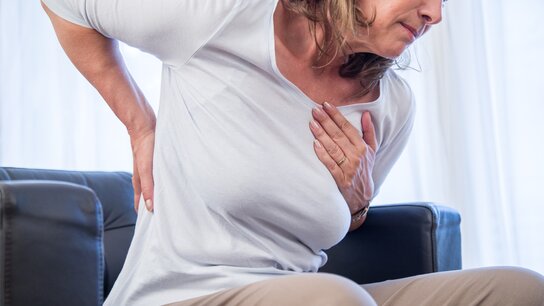 Frau mit Schmerzen im Rücken, sitzt auf einer Couch und hält sich besorgt die Hand an die Brust.
