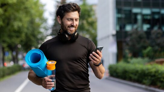 Sportlich gekleideter Mann läuft auf einem Weg, hält ein Handy, eine Yogamatte und eine Wasserflasche, hört Musik.