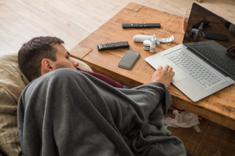 Mann in einer Decke gekuschelt, sitzt an einem Tisch mit Laptop, Fernbedienungen und Handy. Unordnung um ihn herum.
