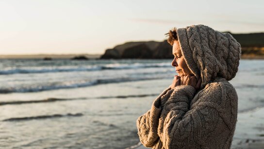 Eine Frau Ende vierzig steht in einen dicken Pullover eingekuschelt am Strand, guckt aufs Meer und wird von der Sonne angeschienen