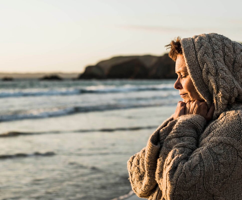 Eine Frau Ende vierzig steht in einen dicken Pullover eingekuschelt am Strand, guckt aufs Meer und wird von der Sonne angeschienen