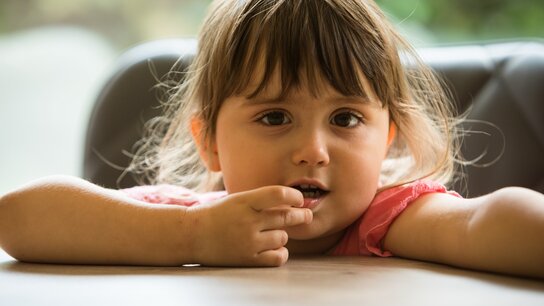 Ein kleines Mädchen mit langen Haaren sitzt an einem Tisch und schaut nachdenklich in die Kamera, während sie nachdenklich mit einem Finger an den Lippen spielt.