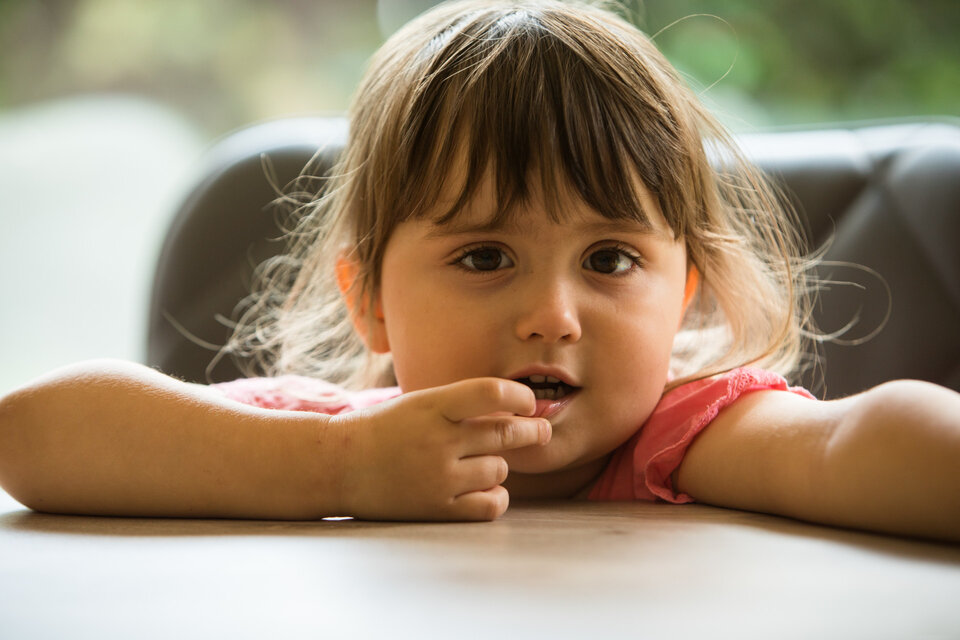 Ein kleines Mädchen mit langen Haaren sitzt an einem Tisch und schaut nachdenklich in die Kamera, während sie nachdenklich mit einem Finger an den Lippen spielt.