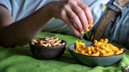 Eine Hand greift nach einer Schüssel mit knusprigen Snacks, während eine andere Schüssel mit Brezeln daneben liegt.