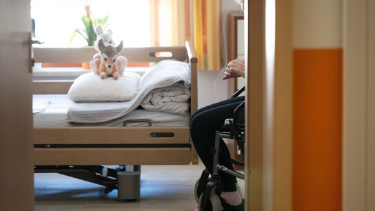 Eine handgestützte Person sitzt auf einem Rollstuhl neben einem Bett, das mit Decken und einem Kuscheltier dekoriert ist.