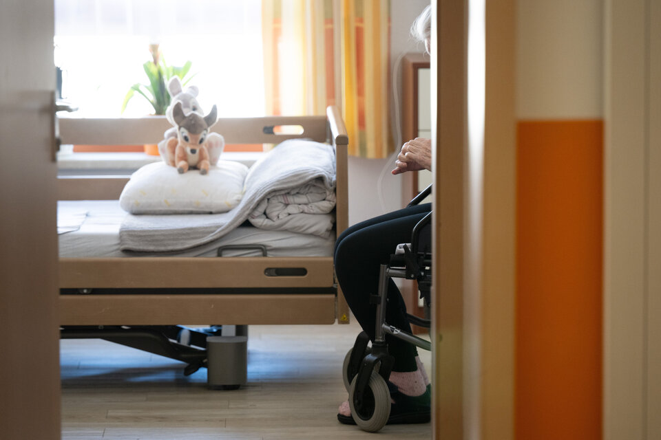 Eine handgestützte Person sitzt auf einem Rollstuhl neben einem Bett, das mit Decken und einem Kuscheltier dekoriert ist.