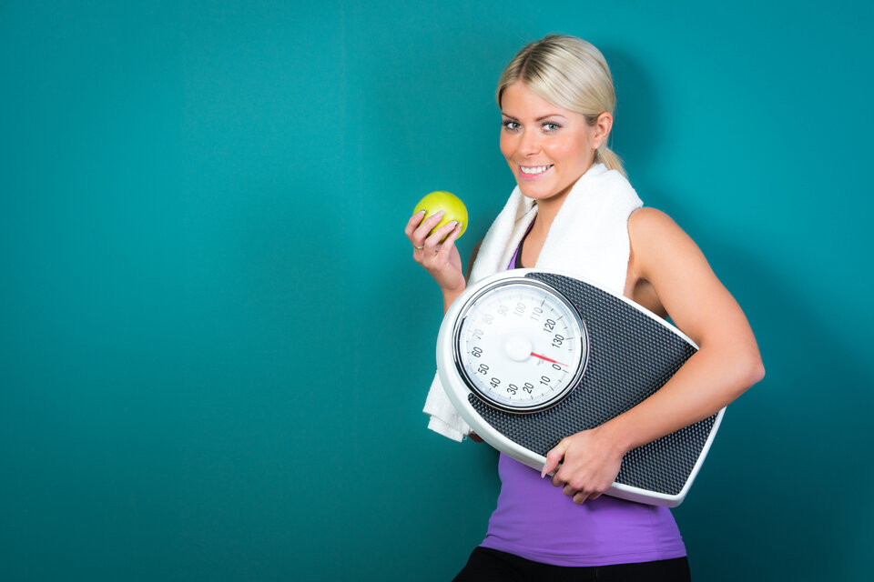 Junge Frau in Sportkleidung hält eine Waage und einen Apfel, mit einem Handtuch um den Hals, vor grünem Hintergrund.