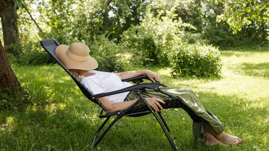 Ein Frau sitzt im grünen Garten im Schatten auf einem Liegestuhl und hat sich einen Strohhut zum Schlafen über das Gesicht gezogen.