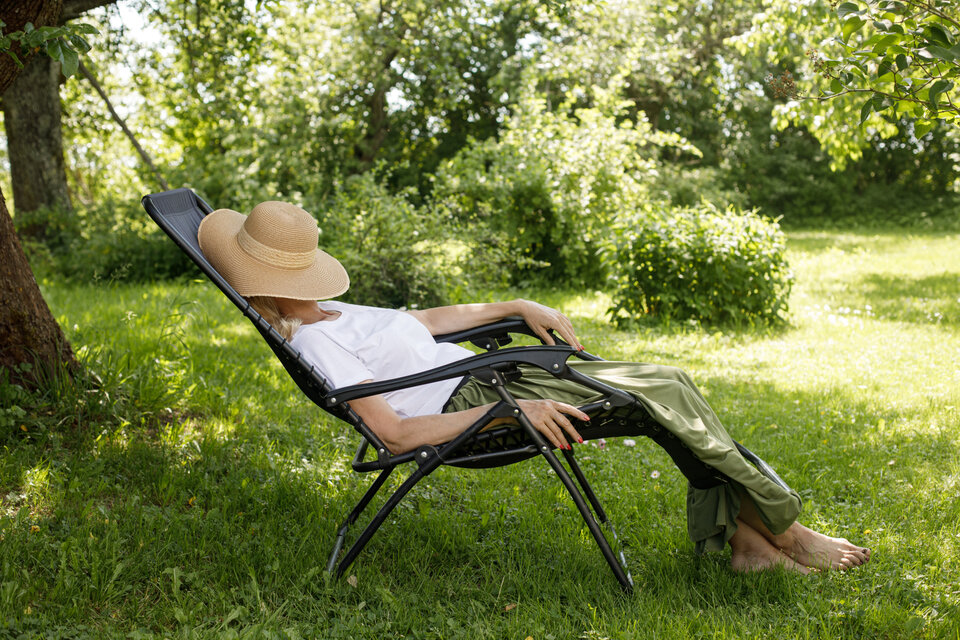 Ein Frau sitzt im grünen Garten im Schatten auf einem Liegestuhl und hat sich einen Strohhut zum Schlafen über das Gesicht gezogen.