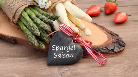 Auf einem Holzbrett liegen ein Bündel weißer Spargel und ein Bündel grüner Spargel. Davor steht ein Schild in Form eines Herzens mit der Aufschrift Spargelzeit.