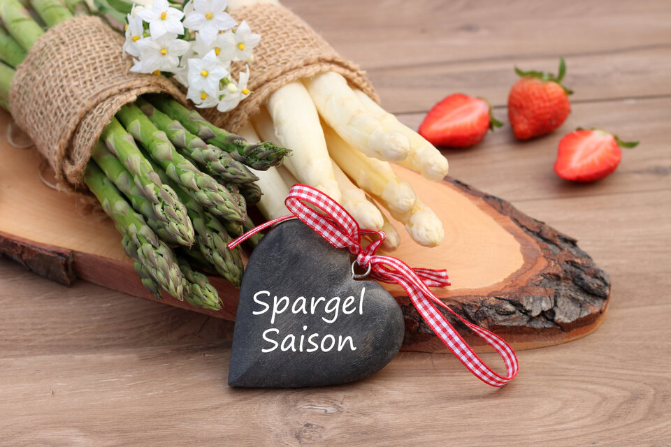 Auf einem Holzbrett liegen ein Bündel weißer Spargel und ein Bündel grüner Spargel. Davor steht ein Schild in Form eines Herzens mit der Aufschrift Spargelzeit.