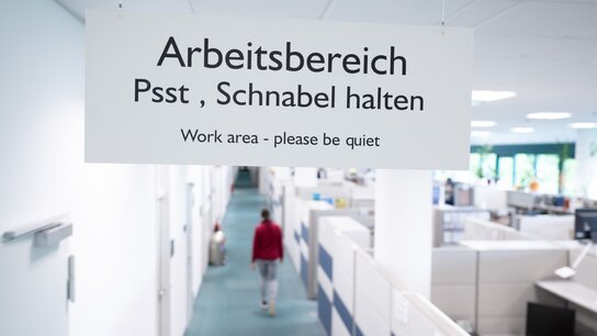 Blick durch eine Tür in ein weiß gehaltenes Großraumbüro. An der Tür hängt ein Schild mit der Auftritt "Arbeitsbereich Psst, Schnabel halten"