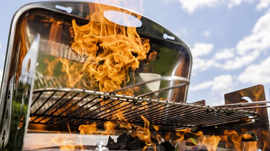 Bei einem Holzkohlegrill schlagen die Flammen sehr hoch. Im Hintergrund ist blauer Himmerl zu sehen.