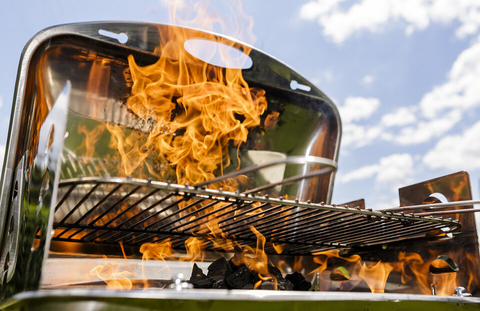 Bei einem Holzkohlegrill schlagen die Flammen sehr hoch. Im Hintergrund ist blauer Himmerl zu sehen.