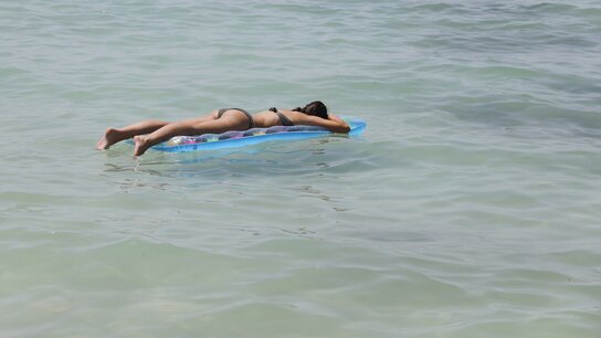 Person in einem schwarzen Bikini liegt auf einem Luftmatratze im seichten Wasser einer Küste.