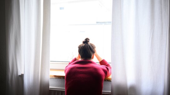 Person sitzt am Fenster, mit den Händen im Nacken, und schaut nach draußen. Helle Vorhänge hängen daneben.