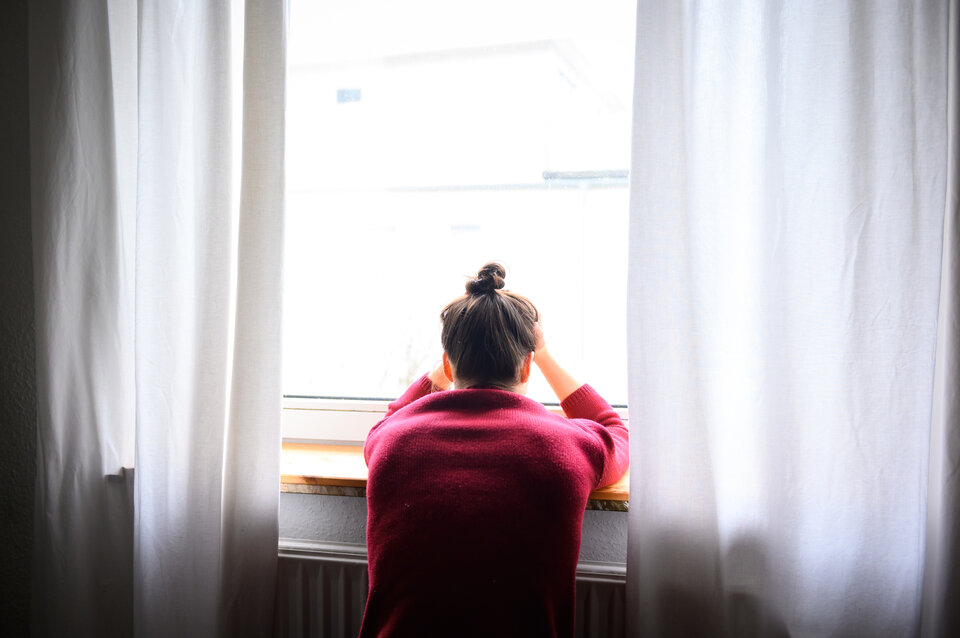 Person sitzt am Fenster, mit den Händen im Nacken, und schaut nach draußen. Helle Vorhänge hängen daneben.