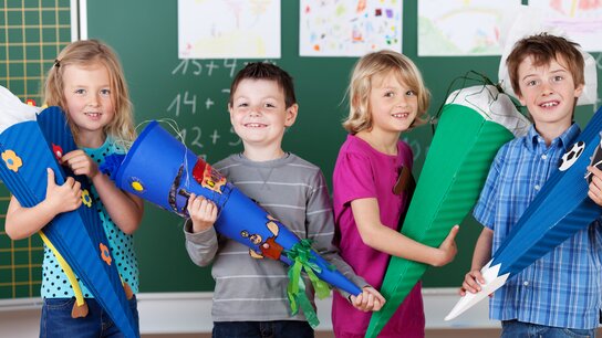 Vier Kinder halten bunte Schultüten in den Händen, lächeln vor einer Tafel mit Zeichnungen im Hintergrund.