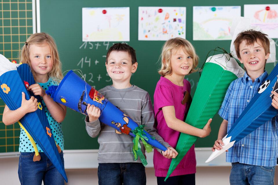 Vier Kinder halten bunte Schultüten in den Händen, lächeln vor einer Tafel mit Zeichnungen im Hintergrund.