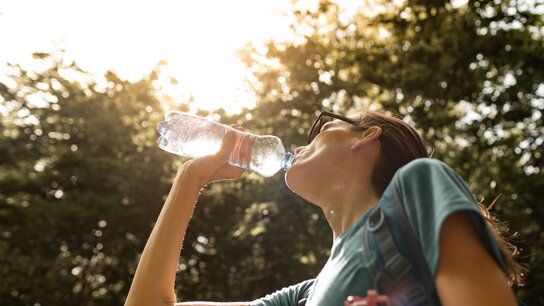 Junge Person trinkt Wasser aus einer Flasche, umgeben von Bäumen, im Sonnenlicht.