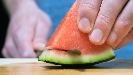 Eine Hand hält ein Stück Wassermelone, während ein Messer das Fruchtfleisch schneidet. Holzbrett im Hintergrund.