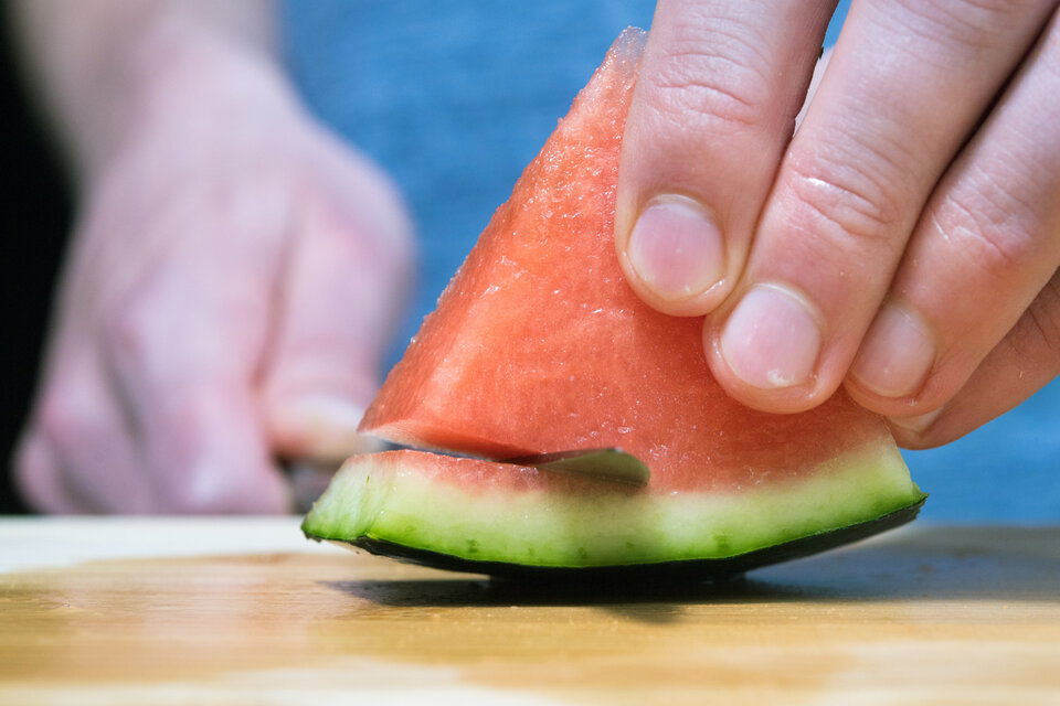 Eine Hand hält ein Stück Wassermelone, während ein Messer das Fruchtfleisch schneidet. Holzbrett im Hintergrund.