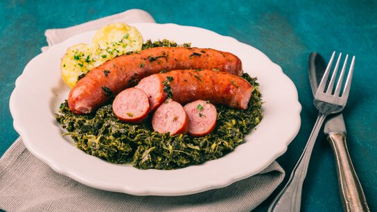 Zwei Bratwürste auf einer Portion Grünkohl, garniert mit Scheiben von Wurst und Beilagen.