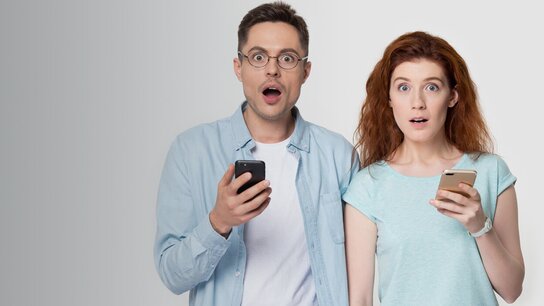 Ein überrascht schauendes Paar hält jeweils ein Smartphone in der Hand, mit offenem Mund und weit aufgerissenen Augen.
