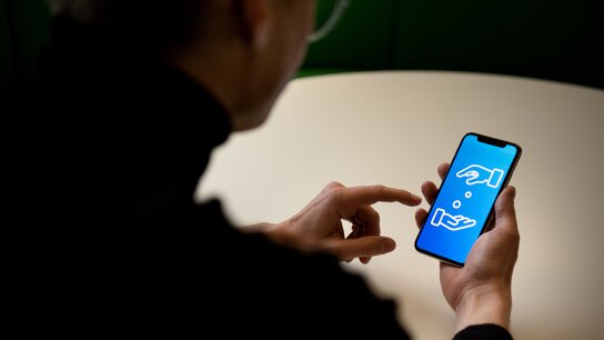 Person hält Smartphone, auf dem ein grafisches Symbol zweier Hände und einer Münze angezeigt wird.