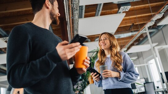 Zwei junge Erwachsene stehen in einem modernen Büro, lächeln und unterhalten sich, während sie Smartphones und Getränke halten.