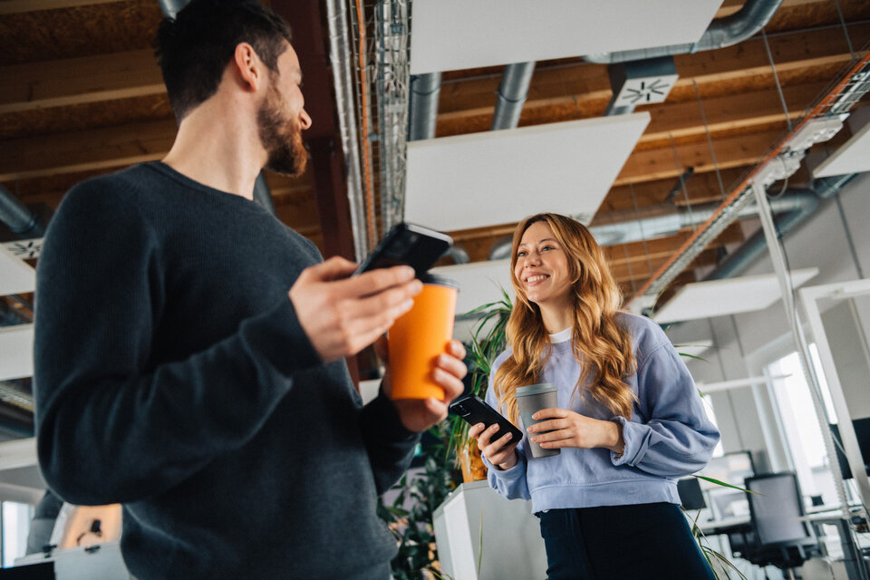 Zwei junge Erwachsene stehen in einem modernen Büro, lächeln und unterhalten sich, während sie Smartphones und Getränke halten.