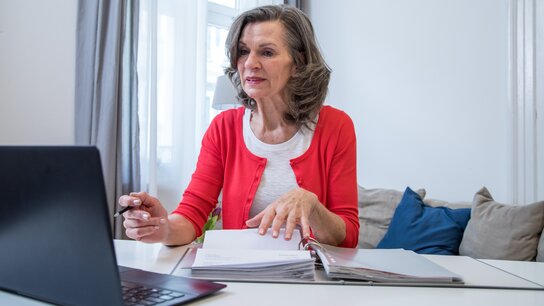 Ältere Frau in rotem Cardigan sitzt an einem Tisch, spricht mit einem Laptop und blättert durch Unterlagen.