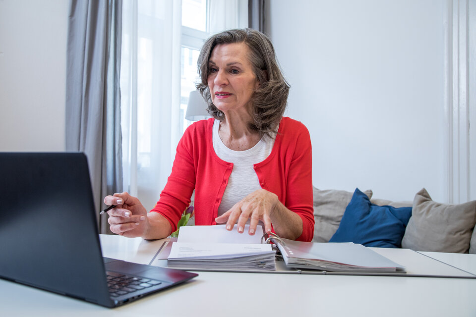 Ältere Frau in rotem Cardigan sitzt an einem Tisch, spricht mit einem Laptop und blättert durch Unterlagen.