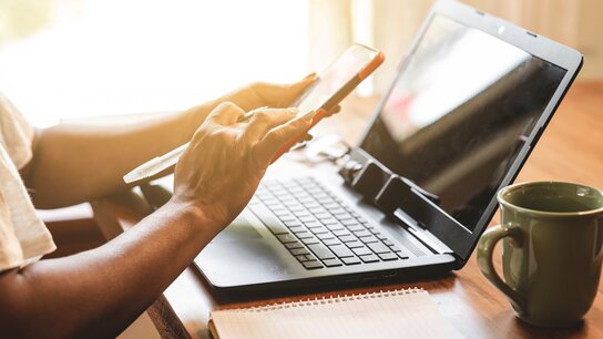 Hände halten ein Smartphone und tippen auf einer Laptop-Tastatur, daneben ein Notizbuch und eine Tasse.