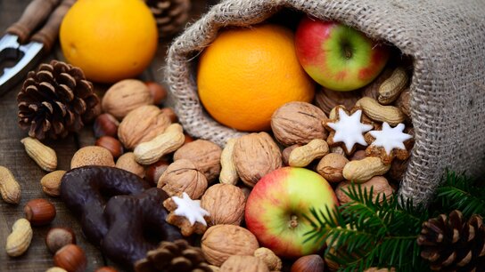 Geschenksack mit Äpfeln, Orangen, Nüssen, Lebkuchen und Tannenzweigen auf einer Holzoberfläche.