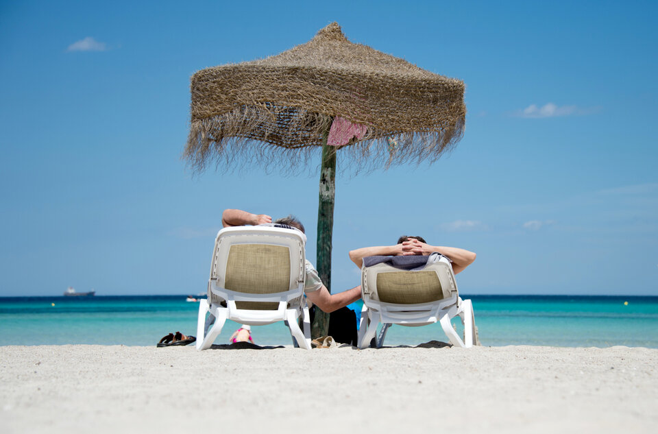 Zwei Personen sitzen unter einem Sonnen-Schirm auf einer Sandstrandliege und genießen den Blick auf das Meer.